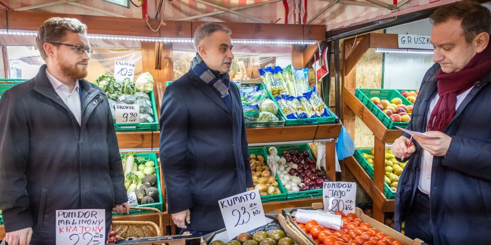 Na zdjęciu trzej mężczyźni rozmawiają na rynku warzywnym, gdzie stoją przed stoiskami z różnymi owocami i warzywami.