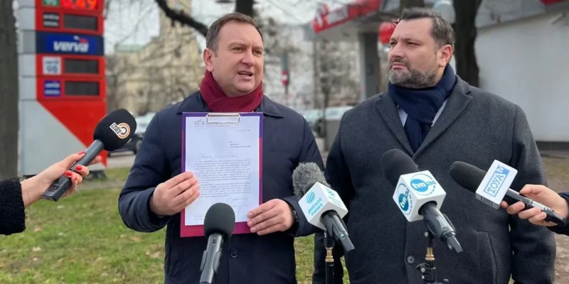 Dwóch mężczyzn w zimowych ubraniach podczas konferencji prasowej na zewnątrz, jeden trzyma klipboard z dokumentem, wokół niego reporterzy z mikrofonami.