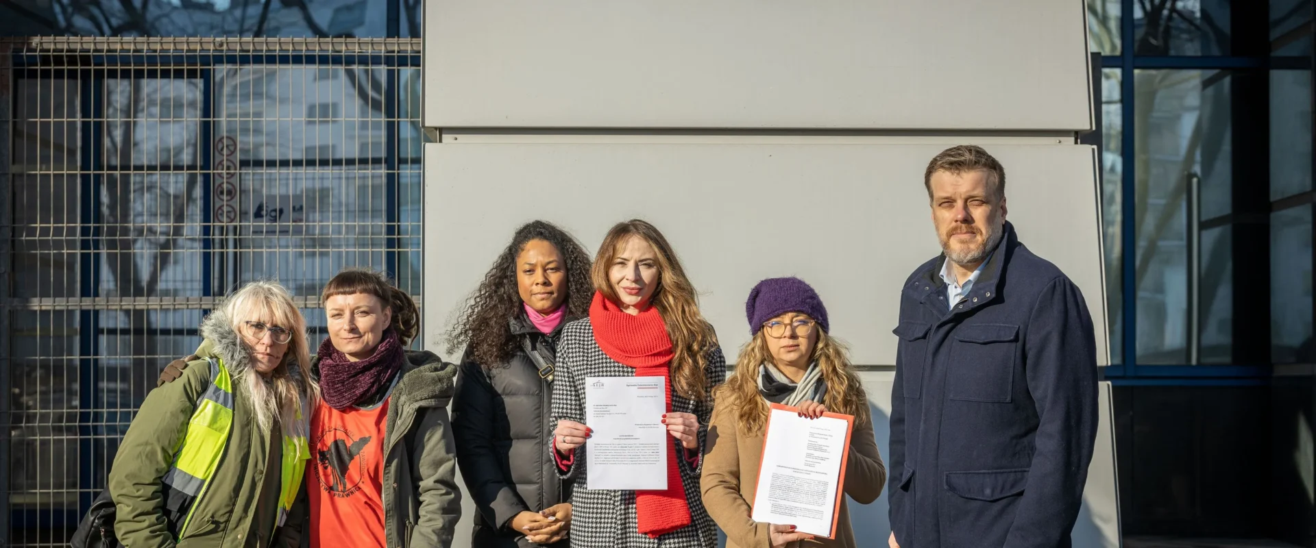 Grupa osób stoi przed budynkiem Prokuratury Krajowej, trzymając dokumenty i prezentując zawiadomienie dotyczące uniemożliwienia przeprowadzenia referendum strajkowego w magazynach Amazon.