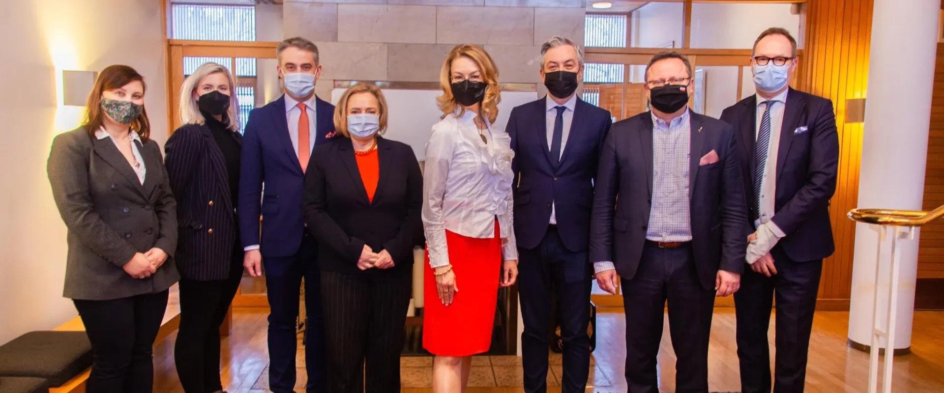 Na zdjęciu znajduje się grupa ośmiu osób, ubranych w eleganckie stroje oraz maseczki, stojących w nowoczesnym wnętrzu.