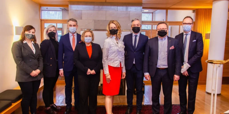 Na zdjęciu znajduje się grupa ośmiu osób, ubranych w eleganckie stroje oraz maseczki, stojących w nowoczesnym wnętrzu.