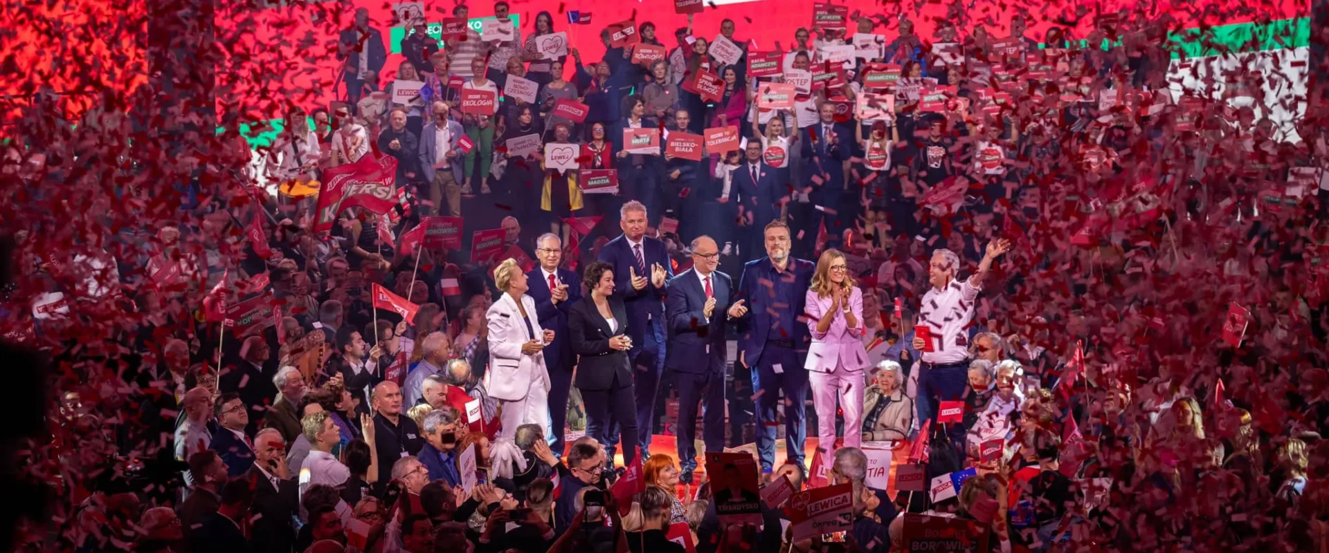 Grupa osób na scenie podczas konwencji w Będzinie, otoczona tłumem i konfetti w kolorze czerwonym, prezentuje hasła związane z hasłem „Każdy głos ma znaczenie”.