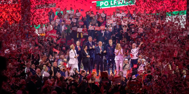 Grupa osób na scenie podczas konwencji w Będzinie, otoczona tłumem i konfetti w kolorze czerwonym, prezentuje hasła związane z hasłem „Każdy głos ma znaczenie”.