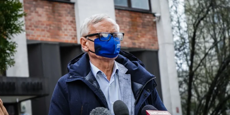 Starszy mężczyzna w okularach i niebieskiej maseczce stoi na zewnątrz przed budynkiem, ubrany w granatową kurtkę i jasnoniebieską koszulę, i przemawia do mediów przy mikrofonach.