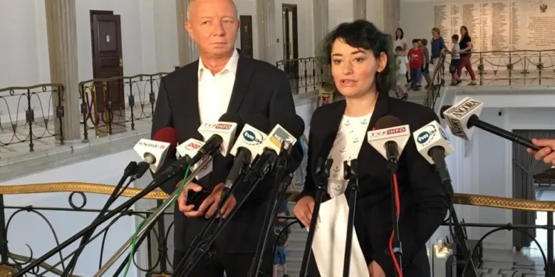 Dwie osoby udzielające wywiadu stoją przed wieloma mikrofonami w reprezentacyjnym wnętrzu, a w tle widać kolumny i przechodzące osoby, w tym dzieci.