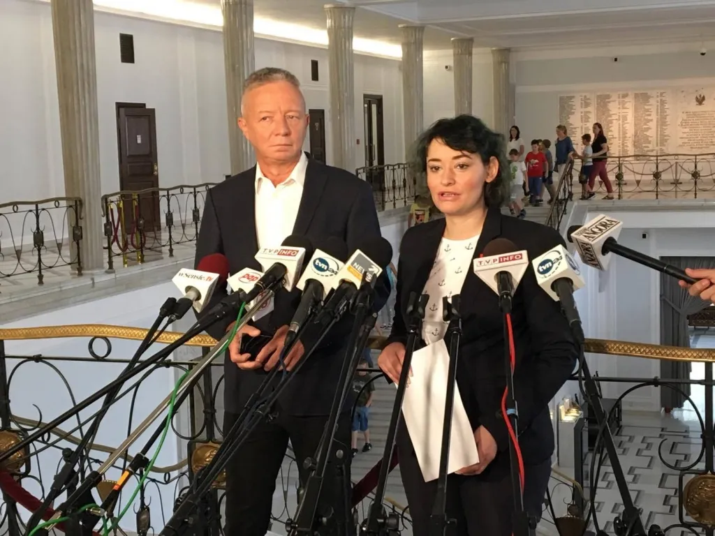 Dwie osoby udzielające wywiadu stoją przed wieloma mikrofonami w reprezentacyjnym wnętrzu, a w tle widać kolumny i przechodzące osoby, w tym dzieci.