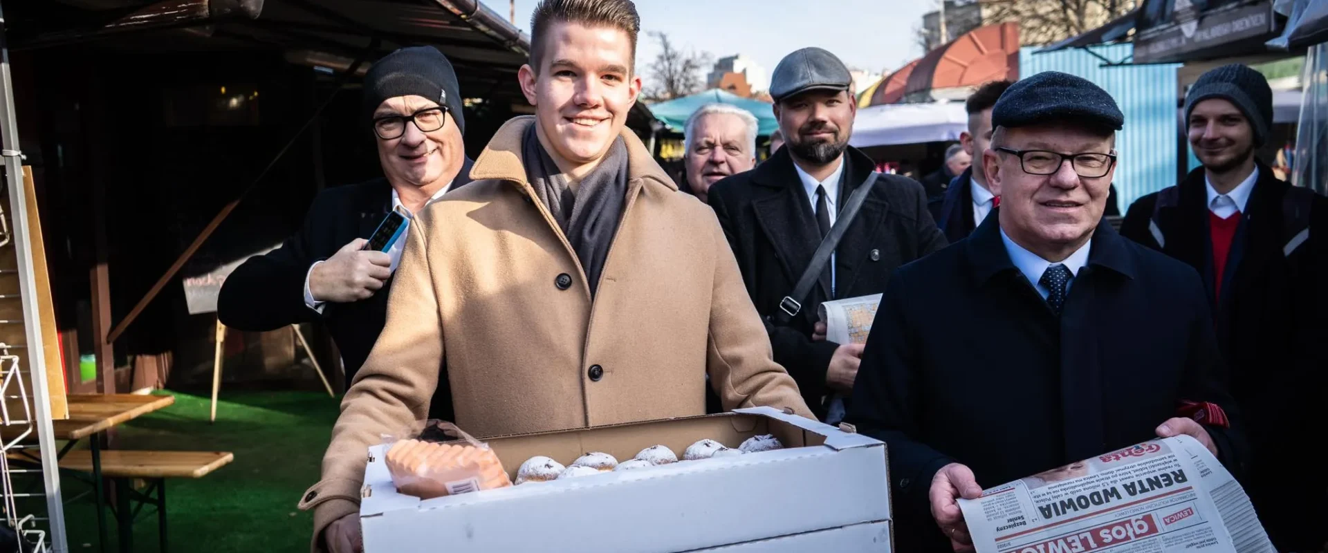 Grupa mężczyzn na targu na Podkarpaciu, na pierwszym planie mężczyzna w beżowym płaszczu trzyma pudełko z jajkami, wokół niego inni uczestnicy i stojące stragany.