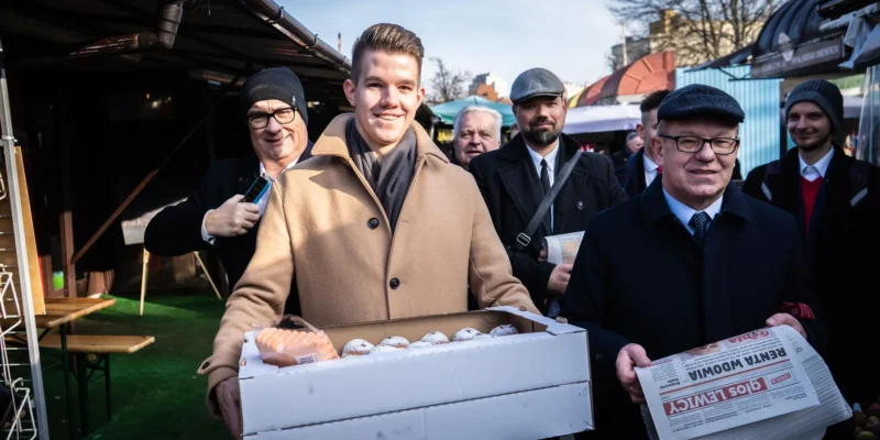 Grupa mężczyzn na targu na Podkarpaciu, na pierwszym planie mężczyzna w beżowym płaszczu trzyma pudełko z jajkami, wokół niego inni uczestnicy i stojące stragany.