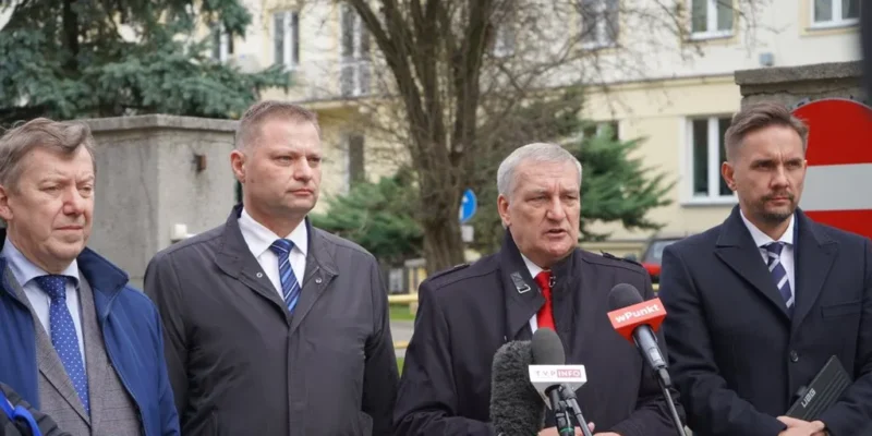 Czterech mężczyzn w formalnych ubraniach stoi przed mikrofonami i kamerami, zdobionych znakami medialnymi, w tle widać budynek i drzewa.