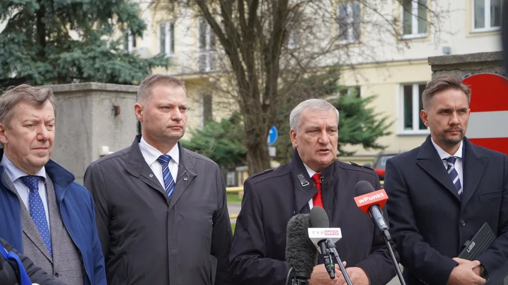 Czterech mężczyzn w formalnych ubraniach stoi przed mikrofonami i kamerami, zdobionych znakami medialnymi, w tle widać budynek i drzewa.