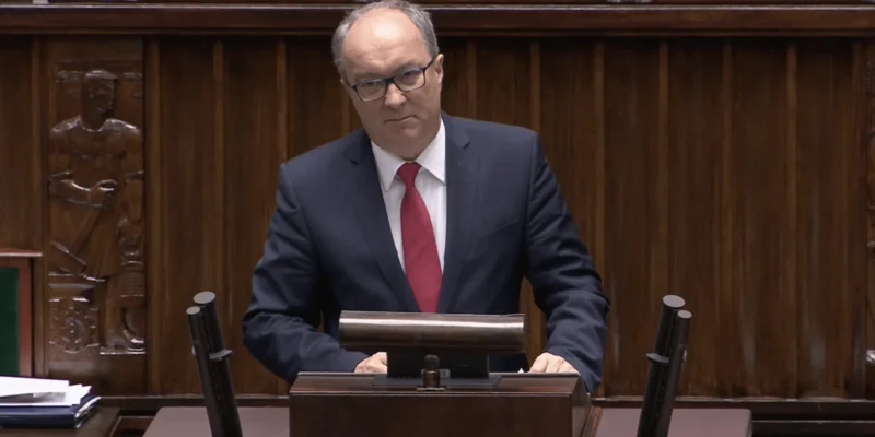 Mężczyzna w granatowym garniturze, czerwonym krawacie i okularach stoi za mównicą w sali plenarnej, na tle drewnianych paneli.