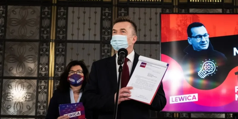 Mężczyzna w garniturze i maseczce przemawia do mikrofonu, trzymając dokumenty, a obok niego stoi kobieta z maseczką z logo Lewicy; w tle widać ekran z kolorową grafiką i napisem Lewica.