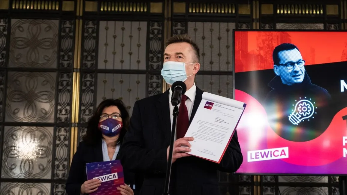 Mężczyzna w garniturze i maseczce przemawia do mikrofonu, trzymając dokumenty, a obok niego stoi kobieta z maseczką z logo Lewicy; w tle widać ekran z kolorową grafiką i napisem Lewica.