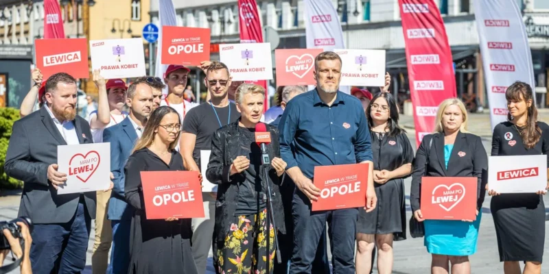 Grupa zwolenników Lewicy na placu w Opolu trzyma plakaty z hasłami „Lewica” i „Prawa Kobiet”, a kobieta przemawia do mikrofonu, domagając się liberalizacji przepisów dotyczących aborcji.