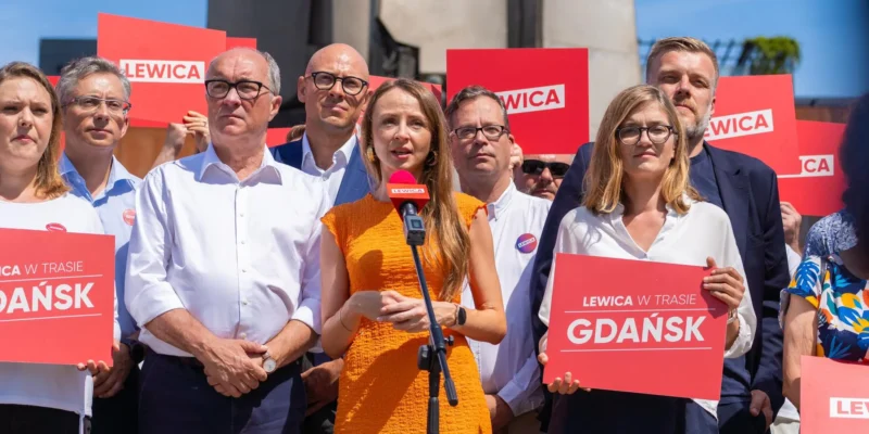 Grupa osób uczestniczy w lewicowej kampanii, trzyma czerwone plakaty z napisem LEWICA, a na środku kobieta w pomarańczowej sukience przemawia do mikrofonu.