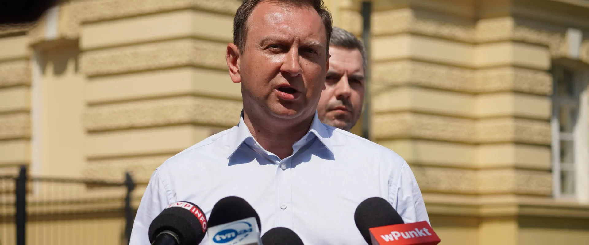 Mężczyzna w białej koszuli przemawia do mediów podczas konferencji prasowej na tle żółtej fasady budynku, stojąc wśród mikrofonów.