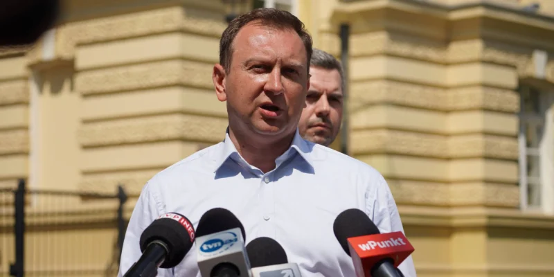 Mężczyzna w białej koszuli przemawia do mediów podczas konferencji prasowej na tle żółtej fasady budynku, stojąc wśród mikrofonów.