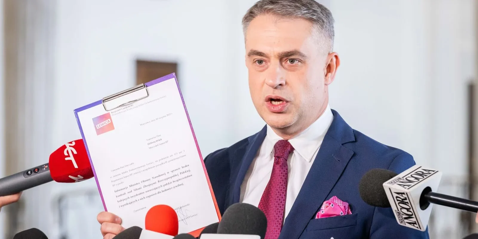 Polityk w granatowym garniturze prezentuje dokument z logo Lewicy podczas konferencji prasowej, otoczony licznymi mikrofonami.