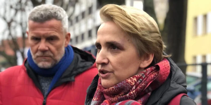 Na zdjęciu widać kobietę w kolorowej chustce mówiącą do kamery, a obok niej stoi mężczyzna w czerwonym płaszczu z poważnym wyrazem twarzy.