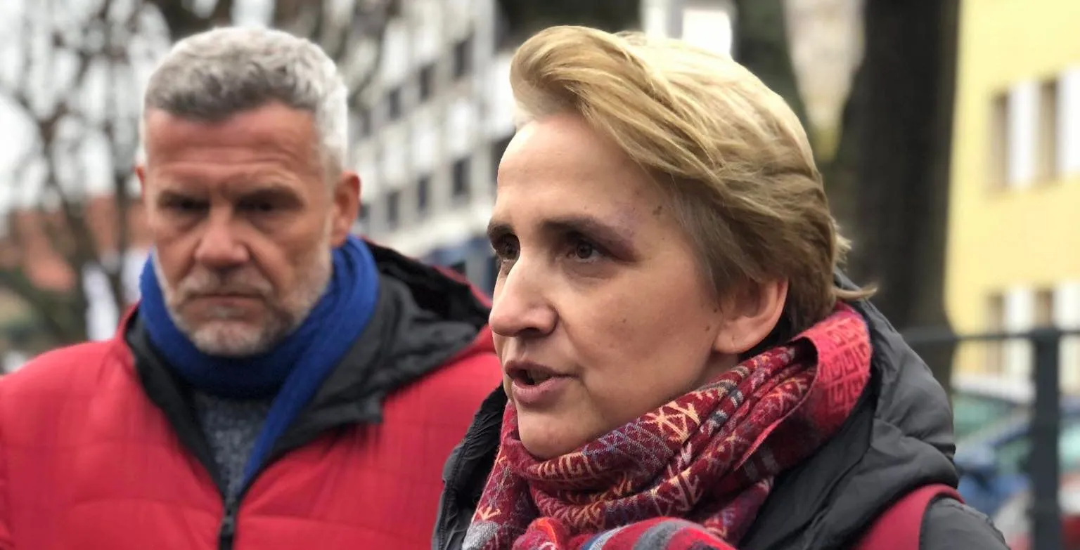 Na zdjęciu widać kobietę w kolorowej chustce mówiącą do kamery, a obok niej stoi mężczyzna w czerwonym płaszczu z poważnym wyrazem twarzy.