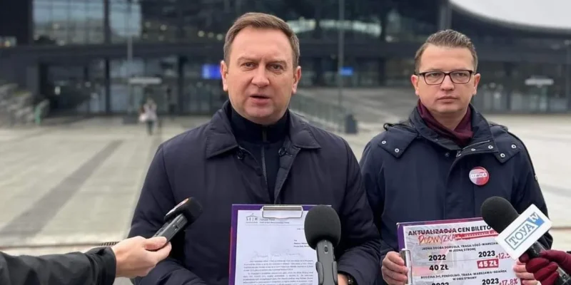 Na zdjęciu widać dwóch mężczyzn w formalnych ubraniach, stojących przed budynkiem stacji Łódź Fabryczna, trzymających dokumenty oraz kartki z informacjami o aktualizacji cen biletów.