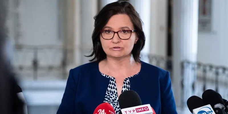 Kobieta w okularach, ubrana w granatową marynarkę, stoi przed mikrofonami podczas konferencji prasowej w jasnym wnętrzu.