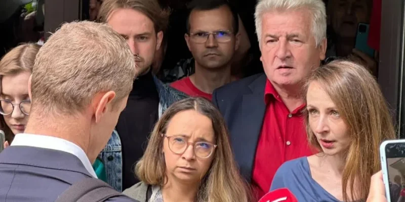 Grupa ludzi stoi przed wejściem do budynku, wśród nich reporterzy z czerwonym mikrofonem, udzielający relacji medialnej.