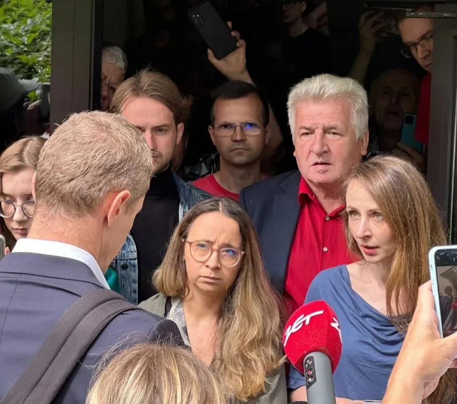 Grupa ludzi stoi przed wejściem do budynku, wśród nich reporterzy z czerwonym mikrofonem, udzielający relacji medialnej.