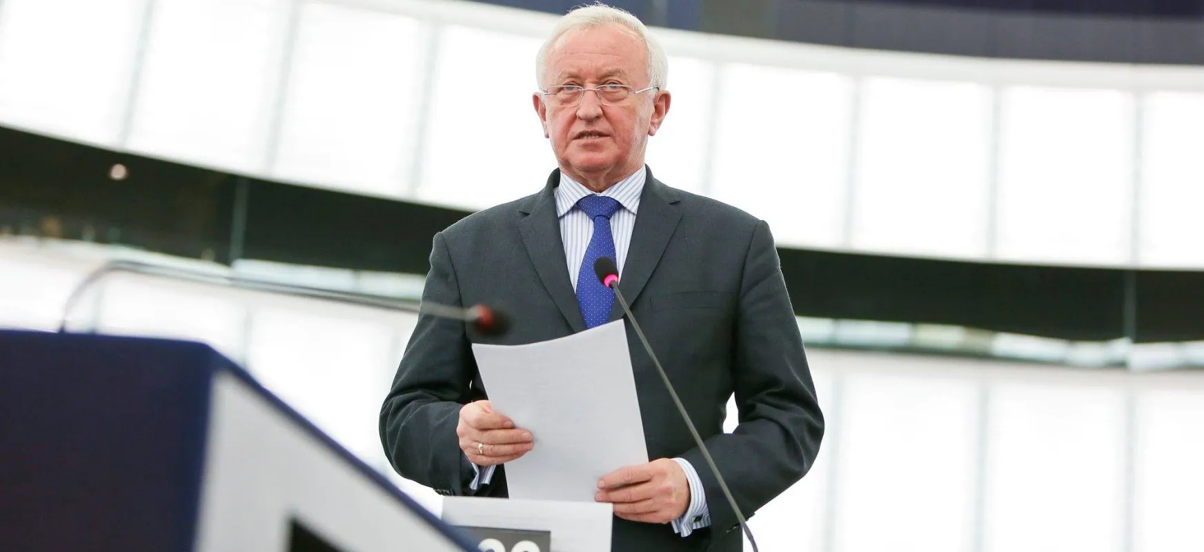 Starszy mężczyzna w garniturze trzyma kartkę papieru i przemawia do mikrofonów podczas konferencji.