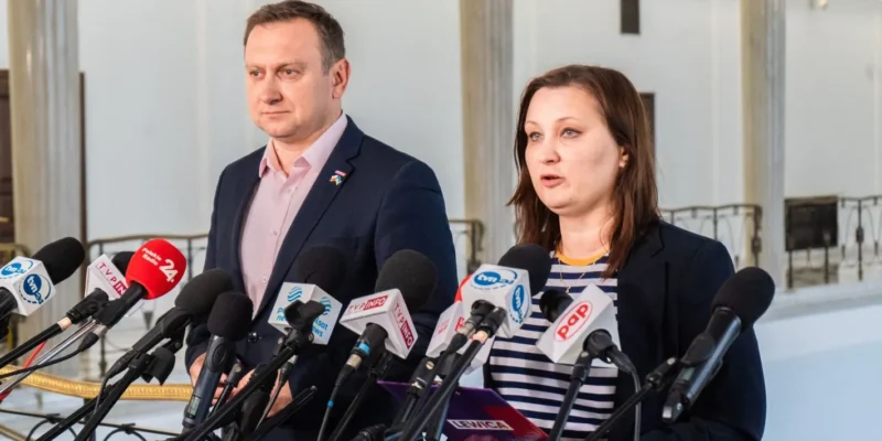 Dwóch przedstawicieli przemawia na konferencji prasowej przed Elektrownią Ostrołęka, otoczonych mikrofonami dziennikarzy.