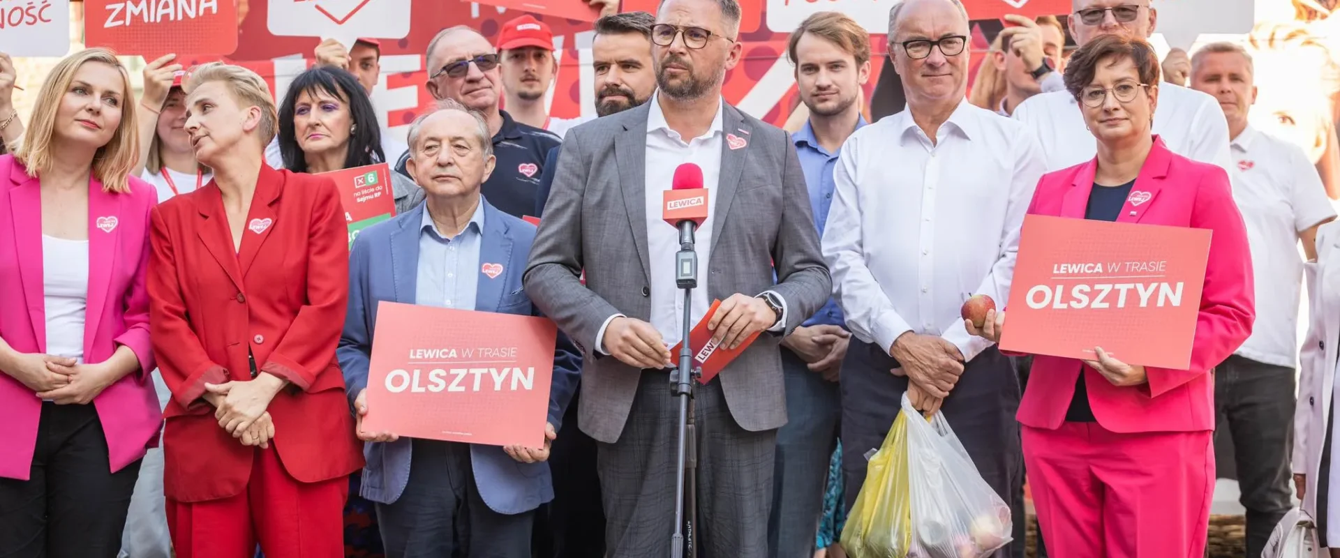 Grupa zwolenników Lewicy na wiecu w Olsztynie, z centralnym mężczyzną w garniturze stojącym przy mikrofonie, trzyma plakaty z napisem „LEWICA W TRASIE OLSZTYN”.
