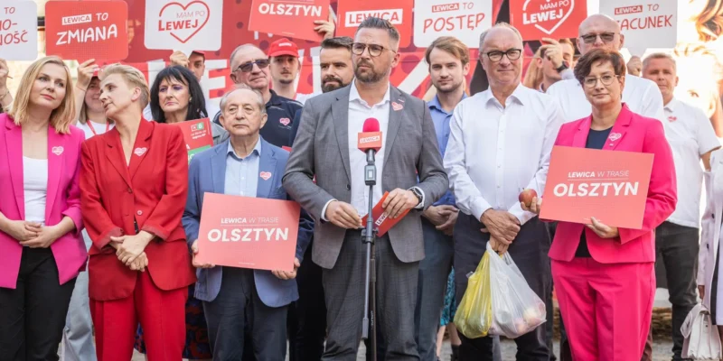 Grupa zwolenników Lewicy na wiecu w Olsztynie, z centralnym mężczyzną w garniturze stojącym przy mikrofonie, trzyma plakaty z napisem „LEWICA W TRASIE OLSZTYN”.