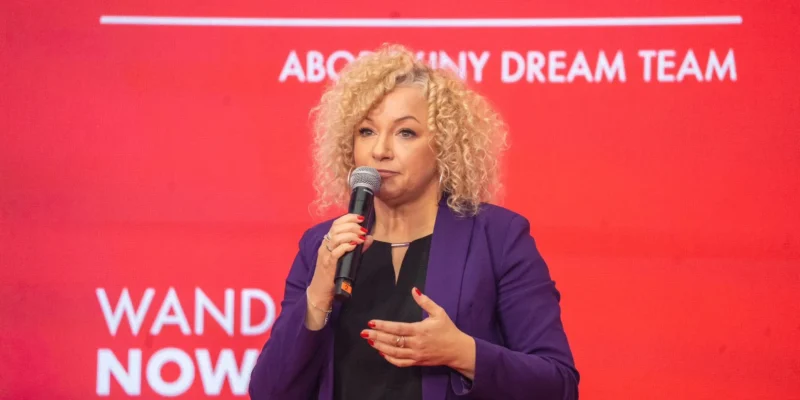 Kobieta o kręconych blond włosach w fioletowym żakiecie przemawia do mikrofonu na czerwonym tle z białymi napisami.