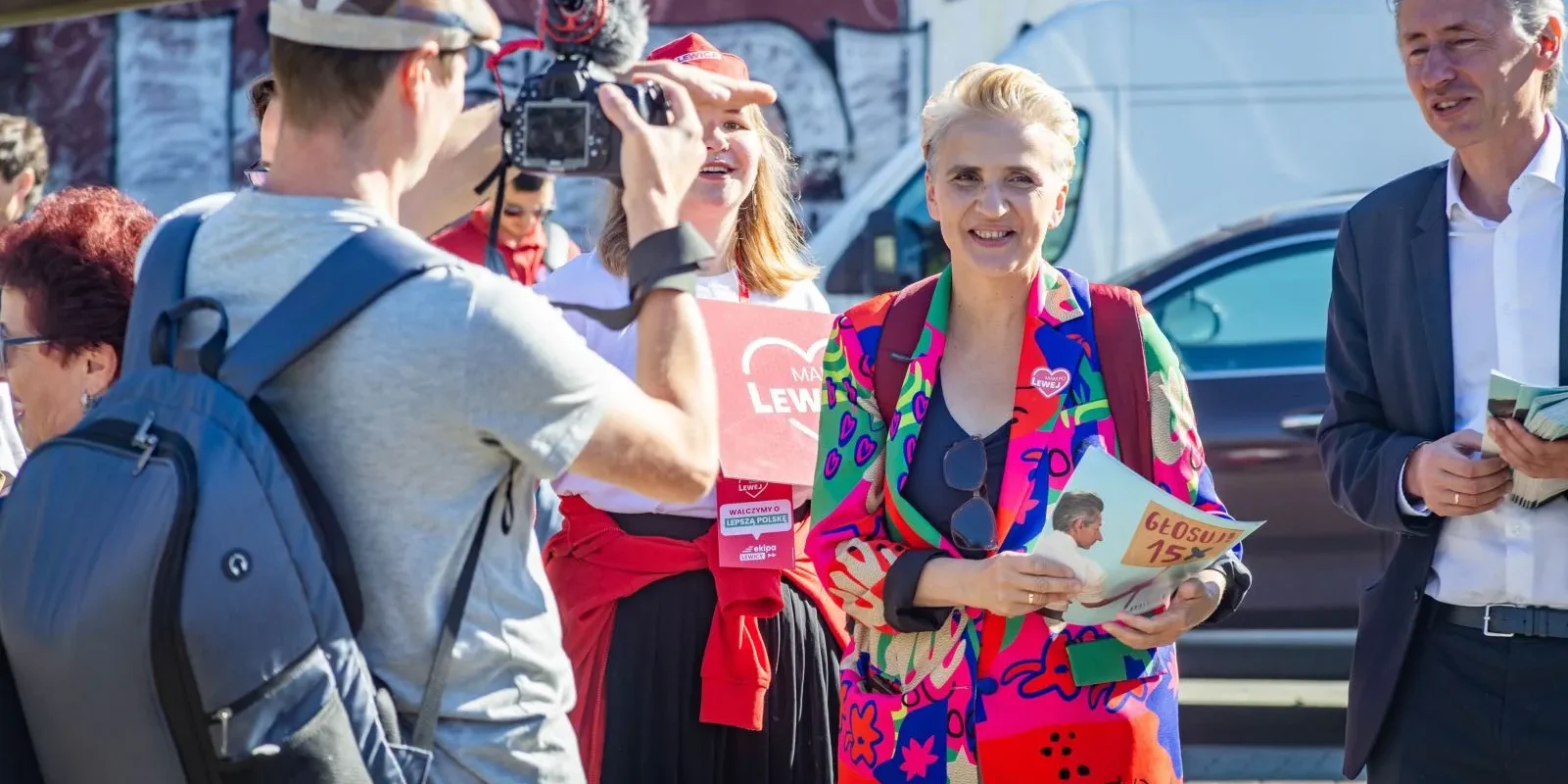 Grupa osób na zewnątrz podczas wydarzenia politycznego; reporter z kamerą fotografuje dwie kobiety i mężczyznę, z których jedna w kolorowym, wzorzystym garniturze trzyma ulotkę i uśmiecha się.