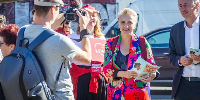 Grupa osób na zewnątrz podczas wydarzenia politycznego; reporter z kamerą fotografuje dwie kobiety i mężczyznę, z których jedna w kolorowym, wzorzystym garniturze trzyma ulotkę i uśmiecha się.