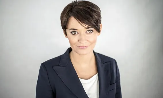 Portret uśmiechniętej kobiety w krótkich włosach, ubranej w ciemną marynarkę i białą bluzkę, na neutralnym jasnym tle.