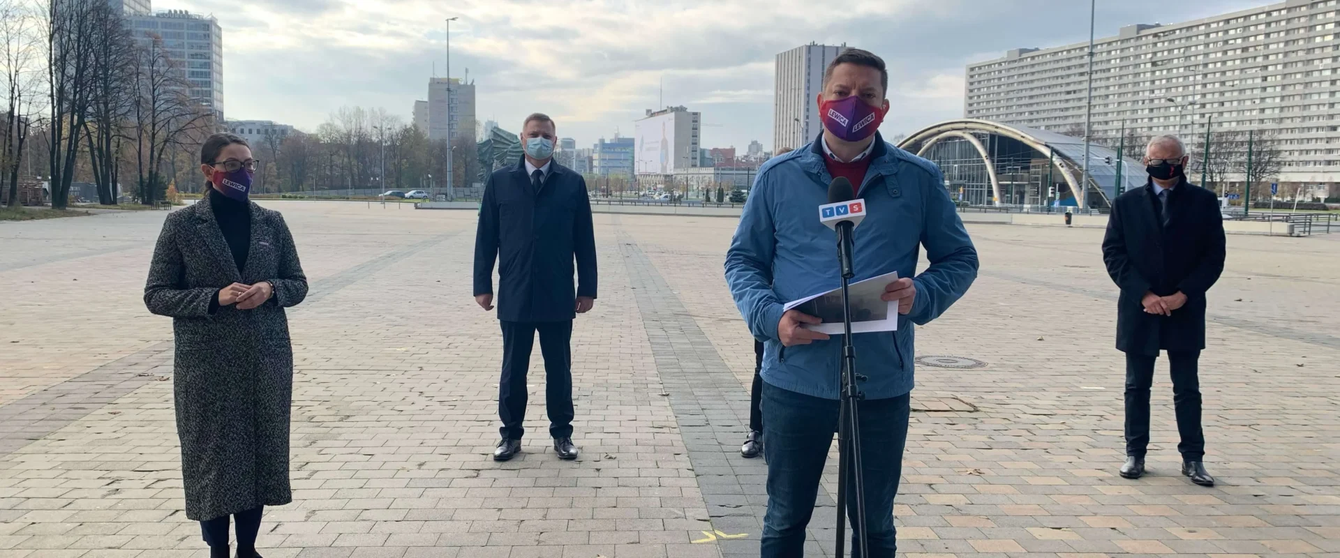 Na otwartym placu czterech ludzi w maseczkach stoi w odstępach podczas konferencji prasowej o walkę ze smogiem w Polsce; mężczyzna na środku mówi do mikrofonu z notatkami, a w tle widać wieżowce.