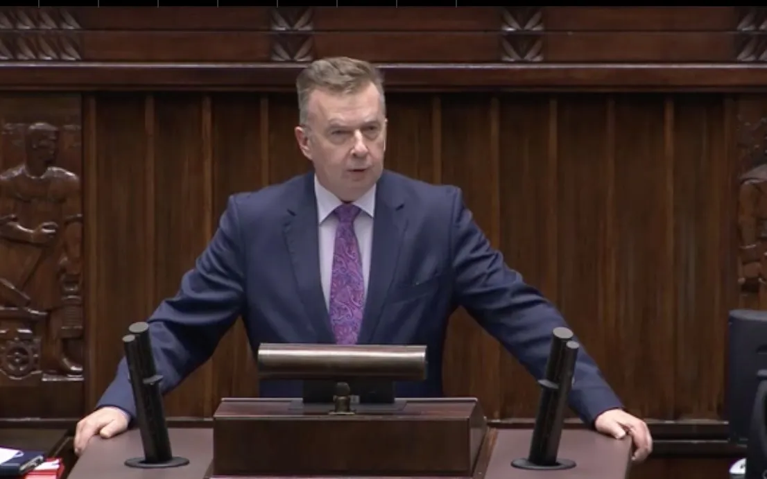 Mężczyzna w granatowym garniturze przemawia zza pulpitu w drewnianej sali plenarnej, stojąc przed mikrofonami.