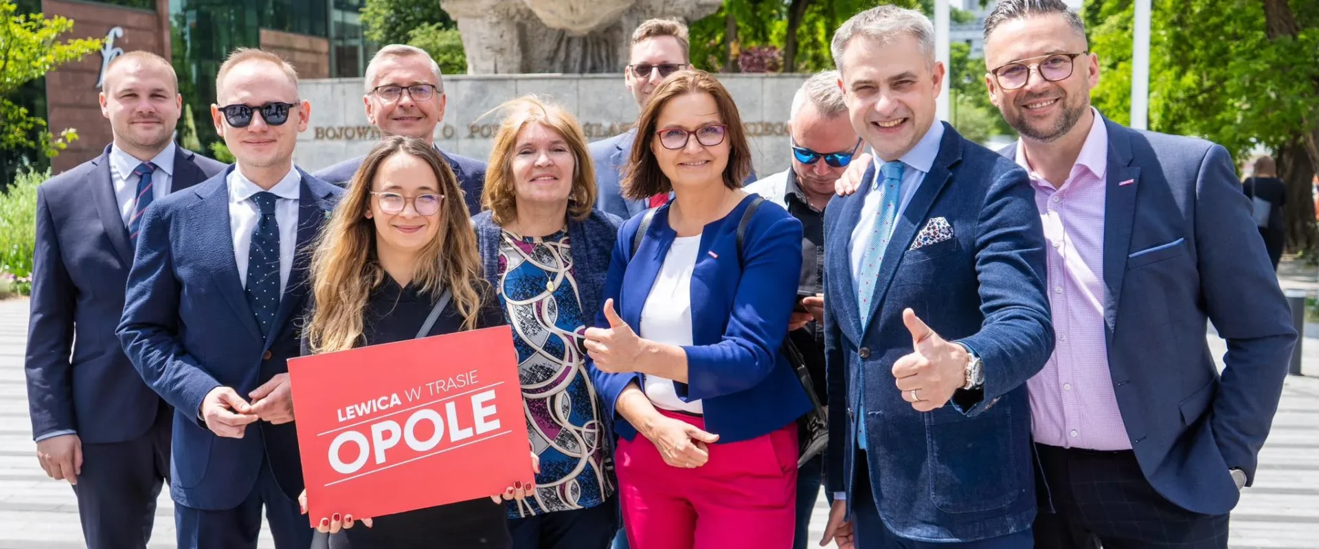 Grupa osób w garniturach stoi przed pomnikiem w Opolu, trzymając czerwony plakat z napisem „LEWICA W TRASIE OPOLE”.