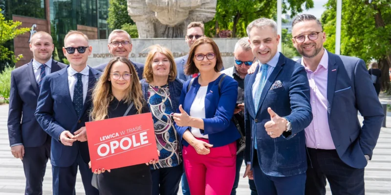 Grupa osób w garniturach stoi przed pomnikiem w Opolu, trzymając czerwony plakat z napisem „LEWICA W TRASIE OPOLE”.