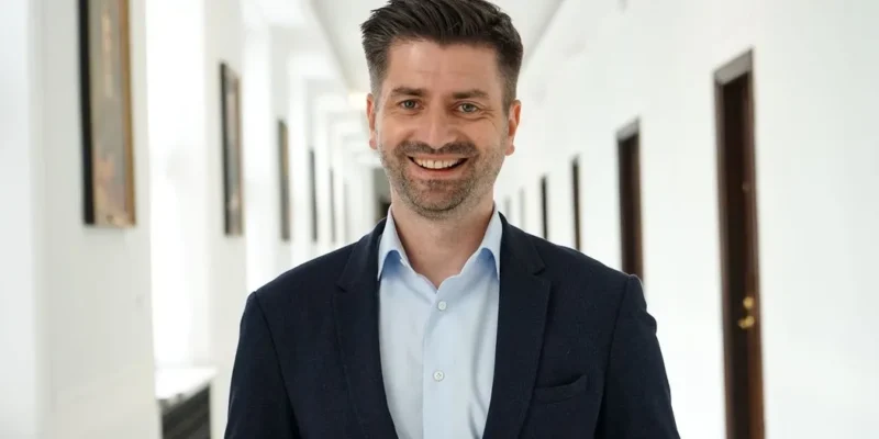 Mężczyzna w garniturze uśmiecha się, stojąc w jasnym korytarzu z zamkniętymi drzwiami po obu stronach.