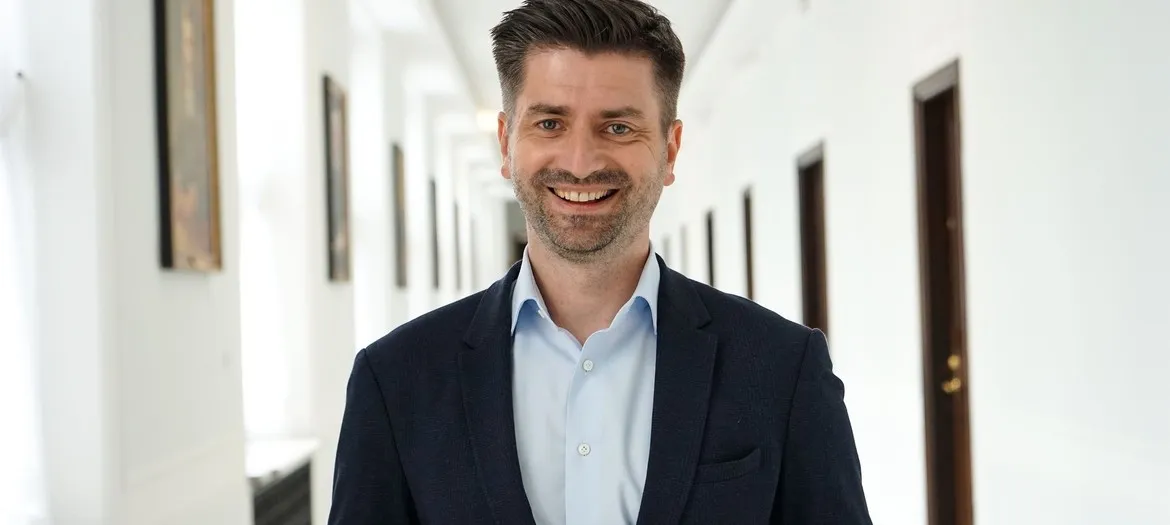 Mężczyzna w garniturze uśmiecha się, stojąc w jasnym korytarzu z zamkniętymi drzwiami po obu stronach.