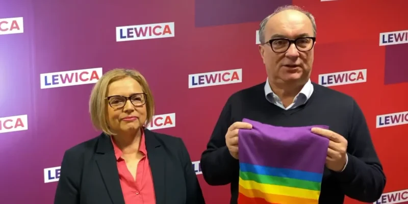 Dwóch ludzi stoi na tle kolorowej ścianki z logotypami Lewicy; mężczyzna trzyma tęczowy komin jako gest wsparcia dla LGBT, wystawiony na aukcję WOŚP.