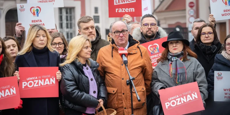 Grupa zwolenników Lewicy na happeningu w Poznaniu trzyma czerwone plakaty „LEWICA w TRASIE POZNAŃ” i gromadzi się wokół mówcy stojącego przy mikrofonie.