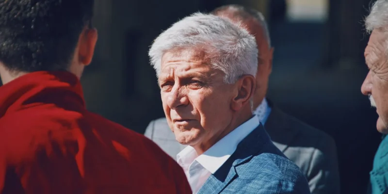 Mężczyzna w niebieskim garniturze i białej koszuli stoi na zewnątrz wśród innych osób, patrząc w bok.