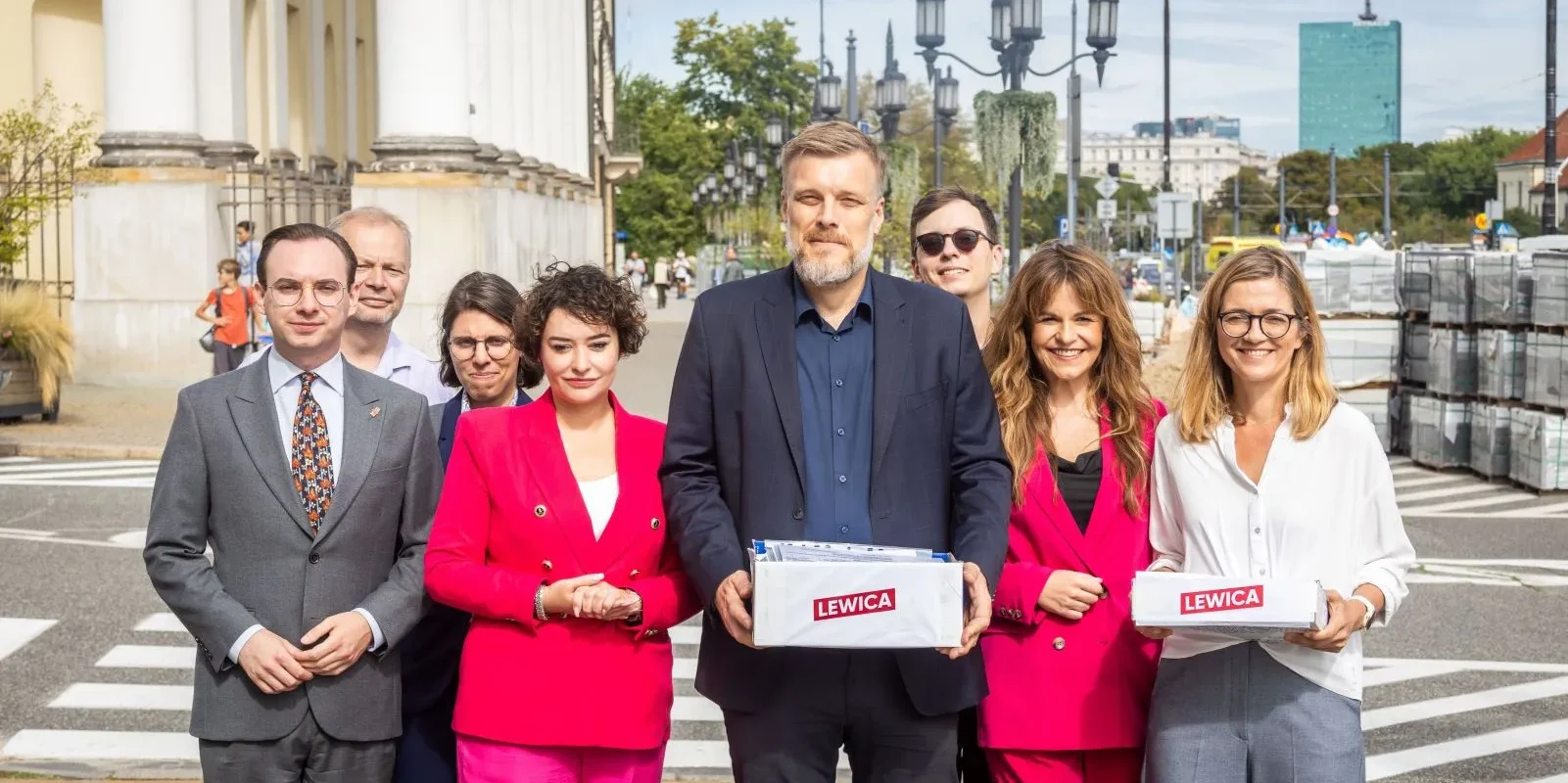 Grupa osób z Lewicy stoi na warszawskim chodniku przy kolumnadzie, trzymając pudełka z logiem Lewica podczas rejestracji list do Sejmu i Senatu.