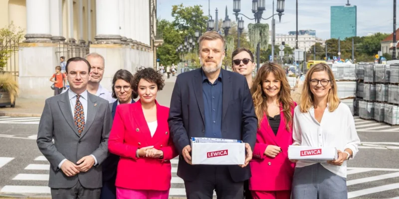 Grupa osób z Lewicy stoi na warszawskim chodniku przy kolumnadzie, trzymając pudełka z logiem Lewica podczas rejestracji list do Sejmu i Senatu.