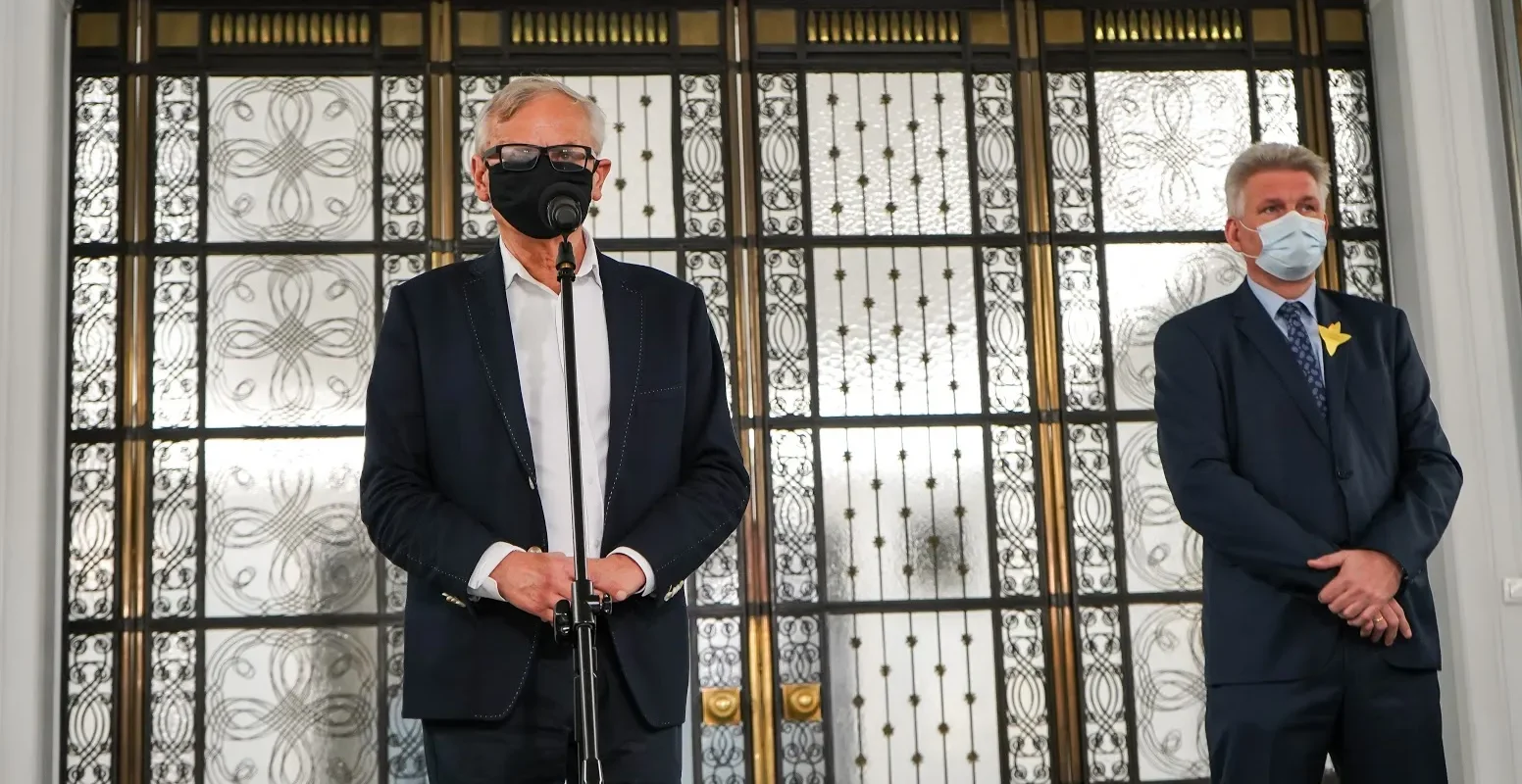 Dwóch mężczyzn w garniturach i maseczkach stoi przy mikrofonie, na tle ozdobnych przeszklonych drzwi z żelaznym ornamentem.