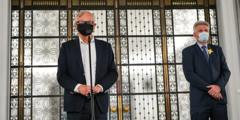 Dwóch mężczyzn w garniturach i maseczkach stoi przy mikrofonie, na tle ozdobnych przeszklonych drzwi z żelaznym ornamentem.