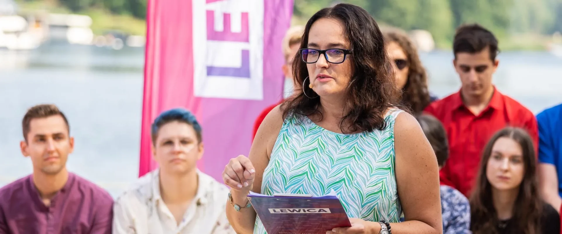 Kobieta w okularach przemawia do zgromadzonych na brzegu jeziora, trzymając ulotkę z napisem Lewica, a w tle widać różowy baner i innych uczestników.